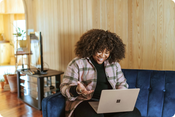 Person sitting on blue couch with laptop, gesturing during online call, with plants and wooden wall in background